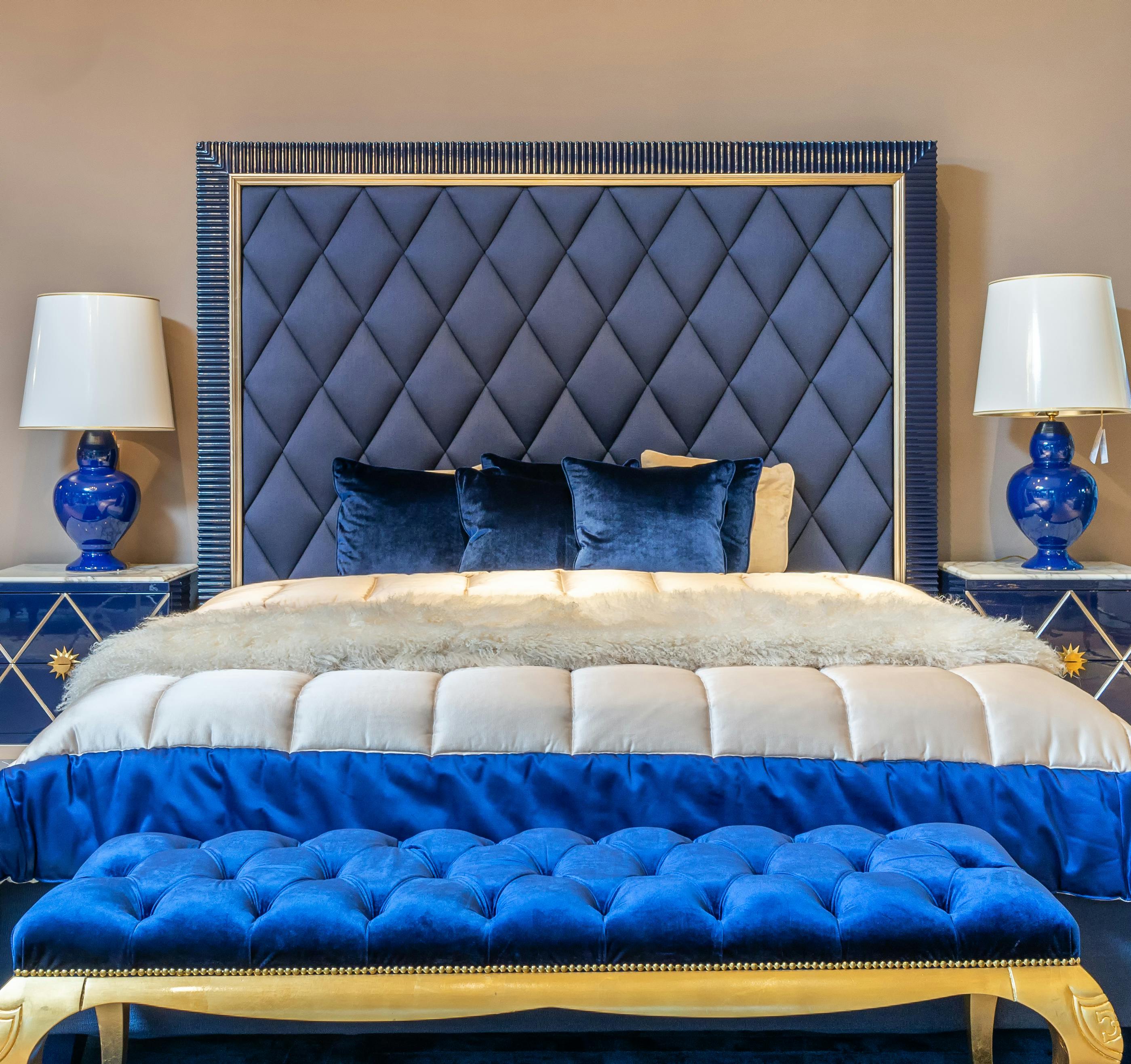 Luxurious blue bedroom featuring a diamond-tufted navy headboard framed in gold, matching velvet bench, cream bedding, and cobalt table lamps on mirrored nightstands.