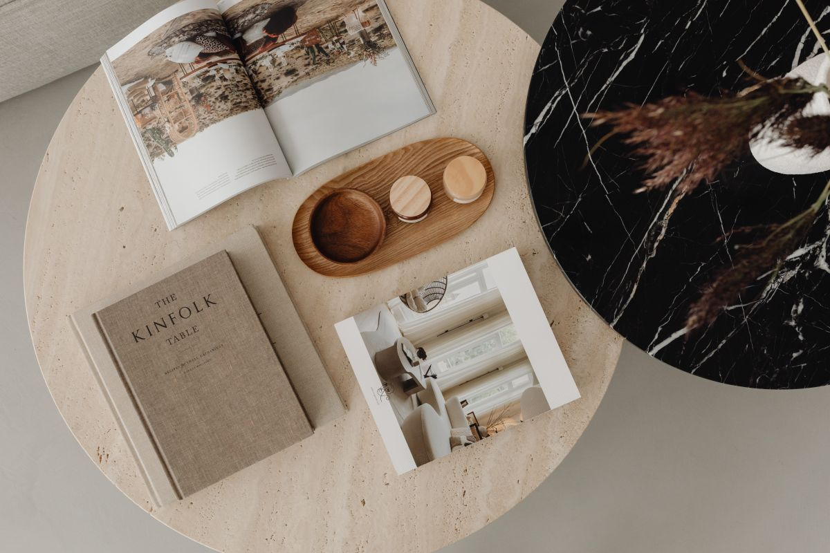 Overhead view of a minimalist living-room vignette: a round travertine coffee table styled with open design magazines, a linen-bound book, and a small wooden tray holding candles, with the edge of a black marble side table and a greige linen sofa visible, all softly lit by natural daylight.”