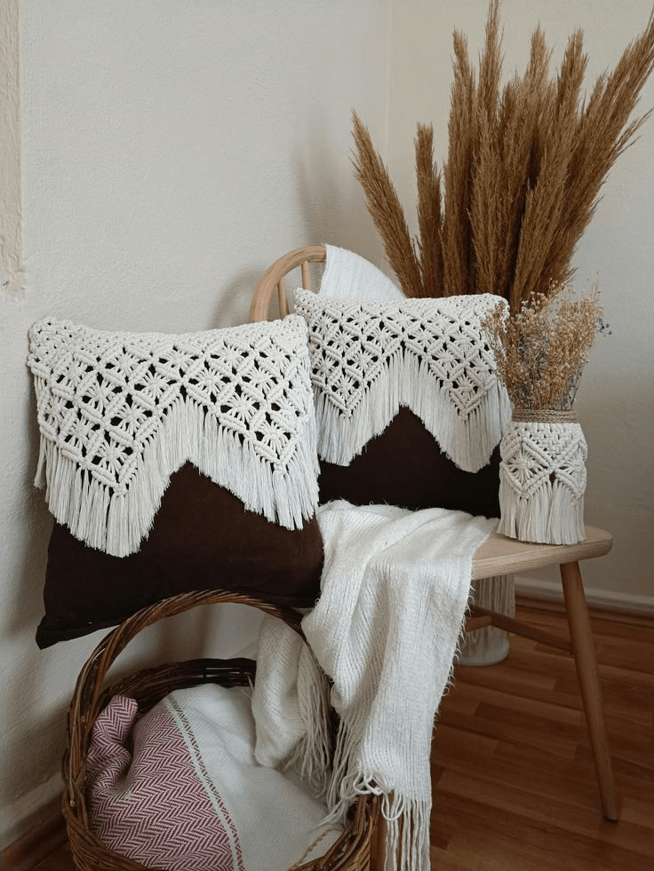 Cozy boho vignette: two square brown throw pillows topped with white macramé crochet fringe rest on a light wooden chair draped in a soft ivory throw; beside them sit a vase of dried pampas grass and a smaller jar of dried florals, while a wicker basket holding folded blankets sits on the warm wood floor beneath.”