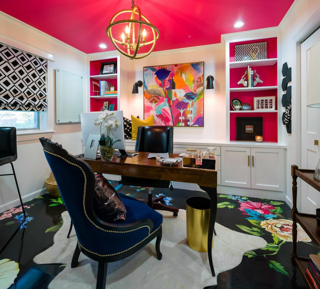 “Home office with bold fuchsia ceiling and built-in white shelving featuring fuchsia back panels, a dark wood desk paired with a navy upholstered chair with nailhead trim, a colorful abstract painting above the desk, geometric black-and-white Roman shade, floral pattern on glossy black flooring, and a modern gold orb chandelier.”
