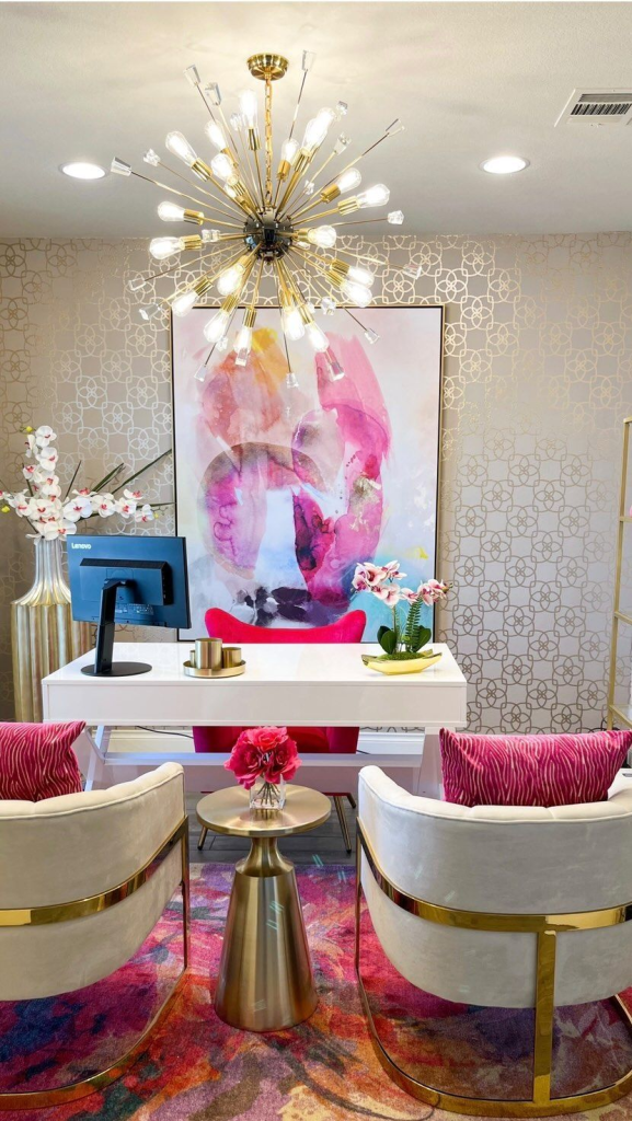 “Chic modern home office with glamorous gold Sputnik chandelier, large abstract pink artwork, white desk with gold accents, velvet white chairs with gold frames, pink zebra-striped cushions, colorful floral rug, and elegant orchid floral arrangements.”