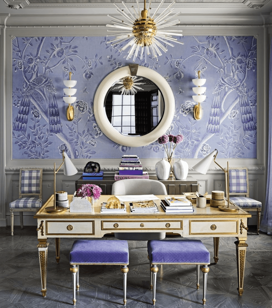 “Elegant home office with ornate gold-trimmed desk, lavender upholstered stools, and a lilac Chinoiserie mural featuring birds and floral motifs, accented by a round wall mirror, gold-and-white sputnik chandelier, gingham chairs, and fresh purple flowers in white vases.”
