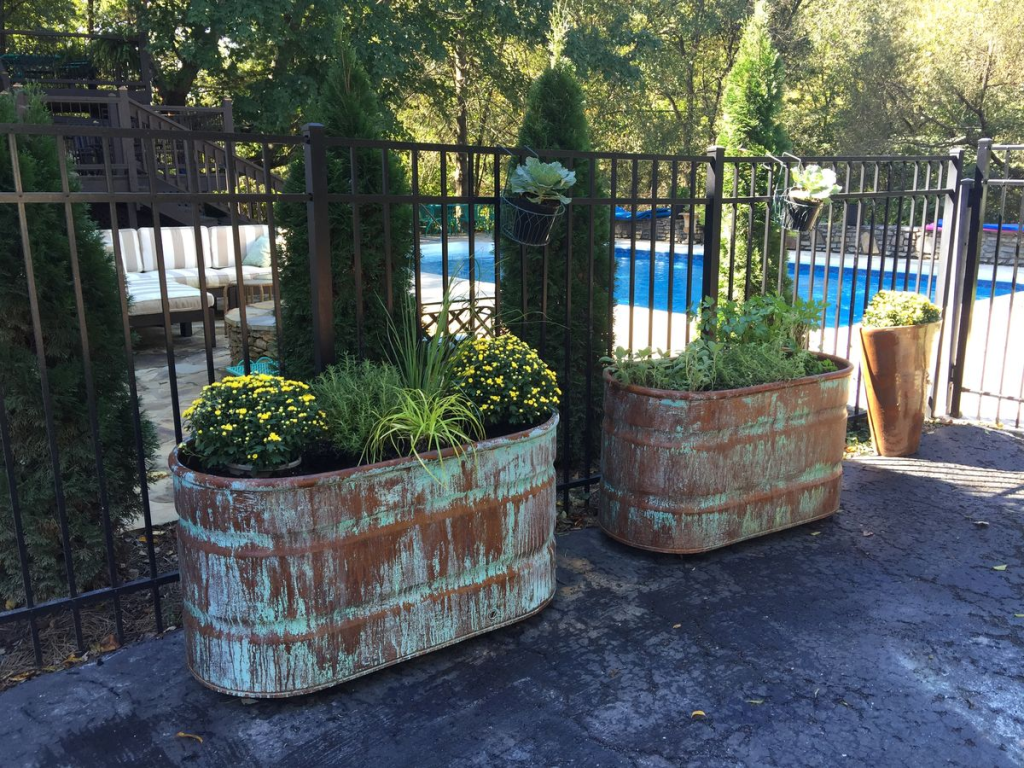 "Outdoor garden planters made from repurposed metal troughs with rustic patina finish, filled with vibrant flowers and greenery by a poolside patio."