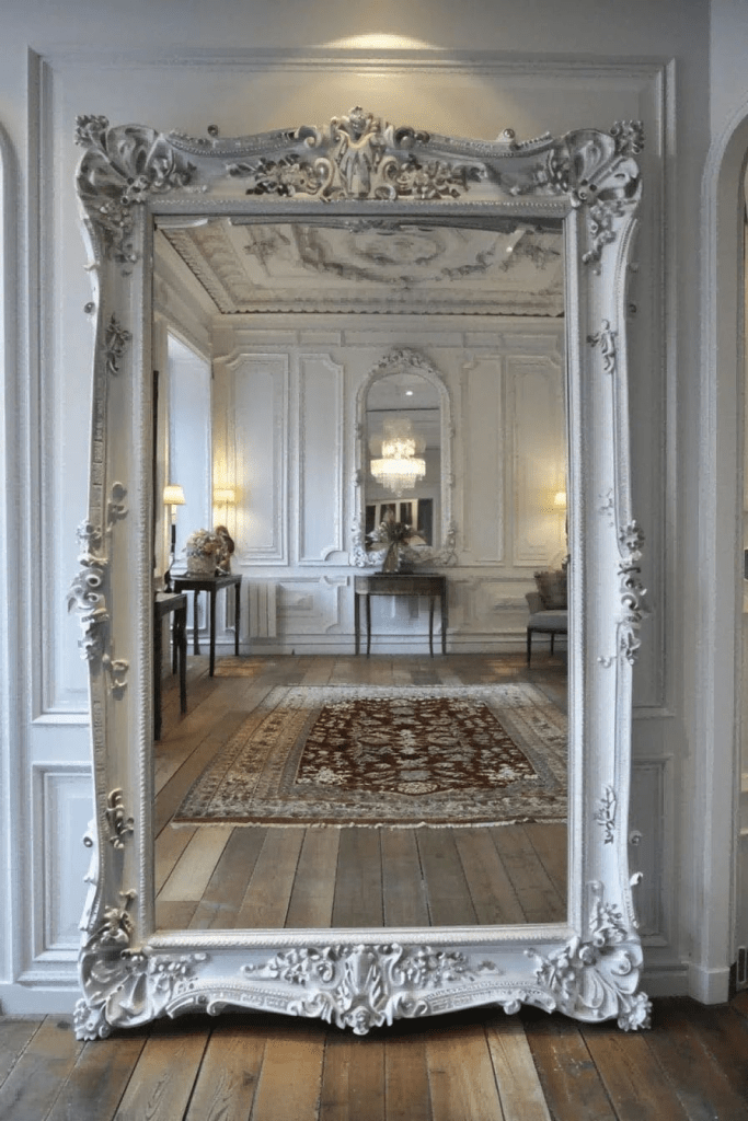 "Elegant ornate full-length mirror with intricate white carved frame reflecting a luxurious French-style interior with chandelier, panelled walls, and Persian rug."