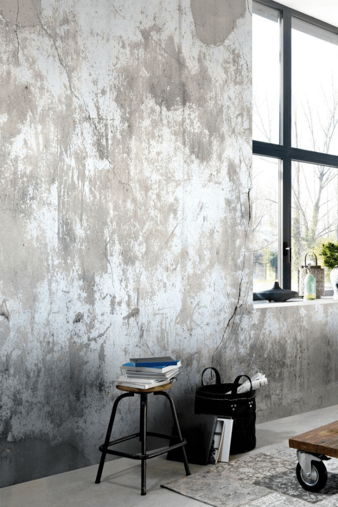 "Modern industrial living space with distressed concrete wall, large black-framed window, vintage stool with books, and rustic coffee table on wheels."