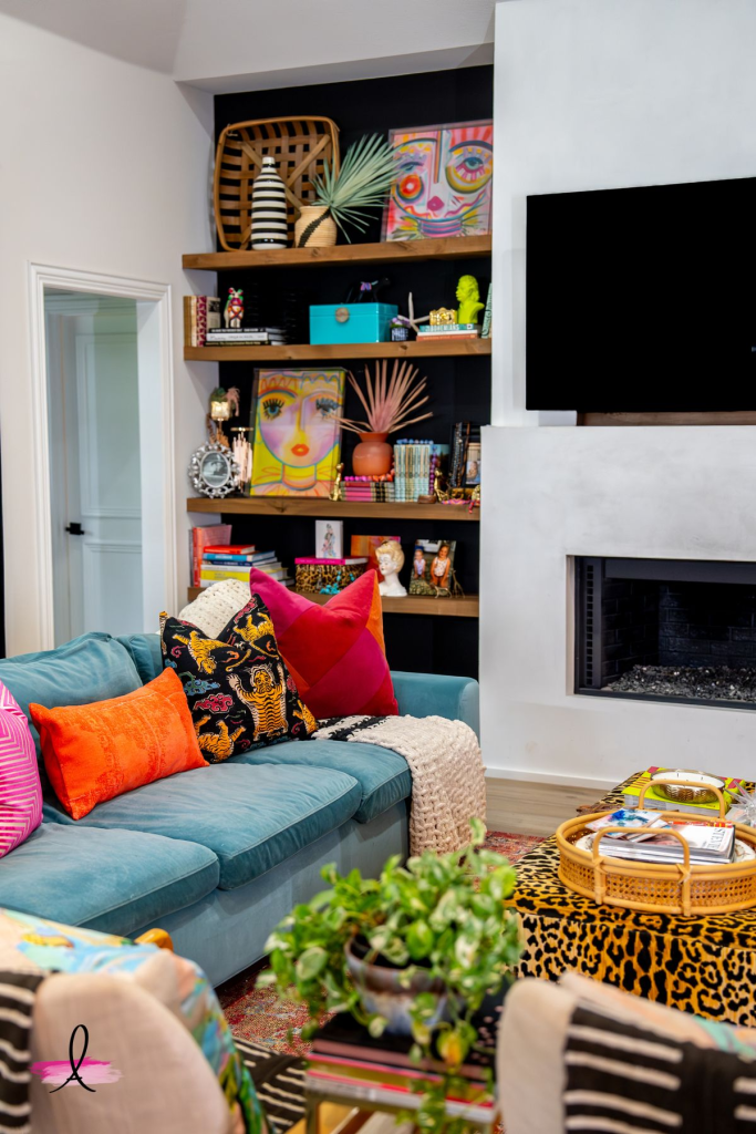 "Eclectic living room with teal sofa, colorful throw pillows, and bold wall art. Playful interior design featuring open wooden shelves styled with books and decor, modern fireplace, leopard print ottoman, and layered textures for a cozy and artistic home style."