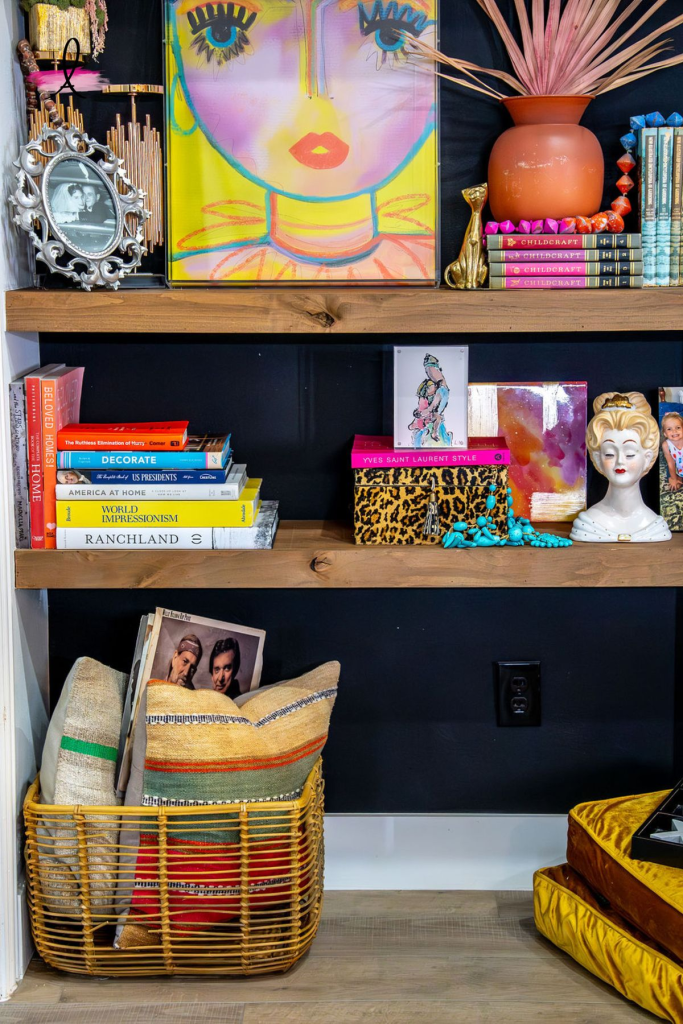 "Eclectic bookshelf styling with colorful books, abstract face art, vintage bust sculpture, and leopard print storage box. Creative home decor featuring wicker basket with pillows, bold accessories, and layered textures for a modern artistic living space."
