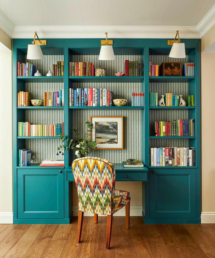 "Chic home office with teal built-in bookshelves, patterned wallpaper backdrop, and colorful book collection. Stylish reading nook featuring a zigzag patterned chair, brass wall sconces, framed artwork, and wooden flooring for a cozy and elegant workspace design."