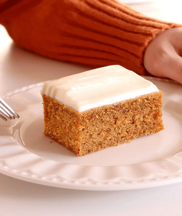 “Moist homemade pumpkin cake slice topped with creamy cream cheese frosting, served on a white plate — a classic fall dessert perfect for cozy autumn baking and Thanksgiving gatherings.”