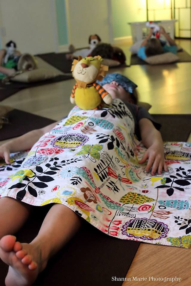 “Child participating in a kids relaxation or mindfulness class, lying on a yoga mat with a patterned blanket and soft plush toy during a guided calm-down or sensory relaxation session.”