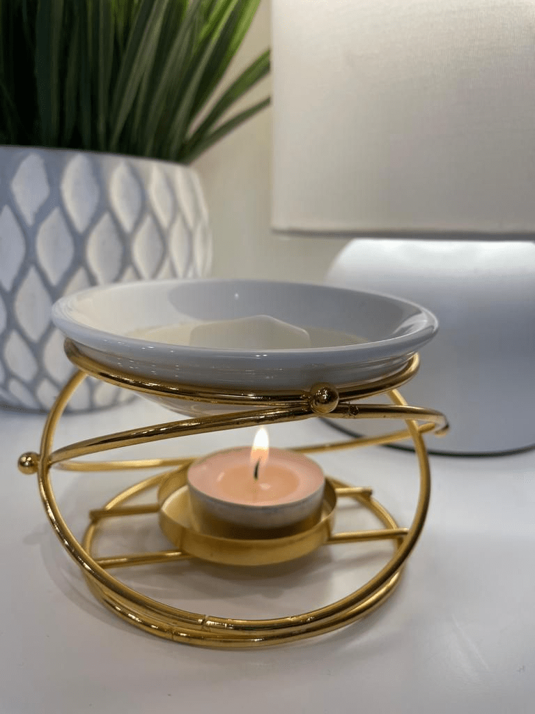 “Elegant gold metal wax melt burner with a lit tealight candle and white ceramic dish, styled on a modern white table beside a plant pot and table lamp, creating a cozy and relaxing home ambiance.”