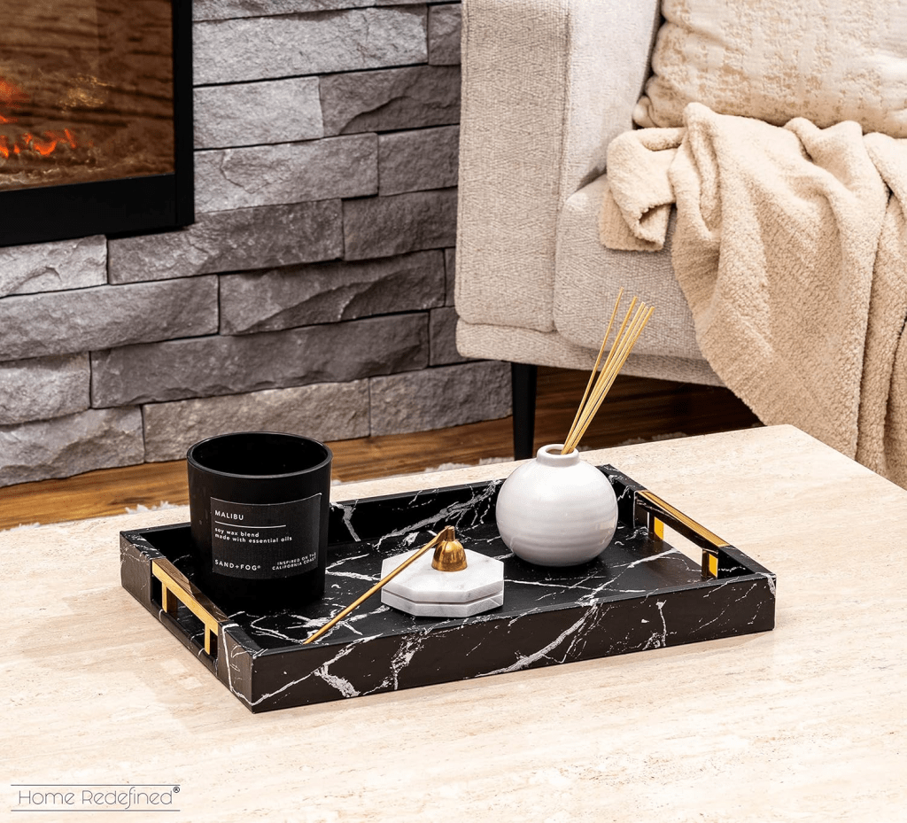 Modern black marble decorative tray with gold handles styled on a coffee table, featuring a scented candle, reed diffuser, and minimalist home accessories in a cozy living room with stone fireplace and neutral decor.