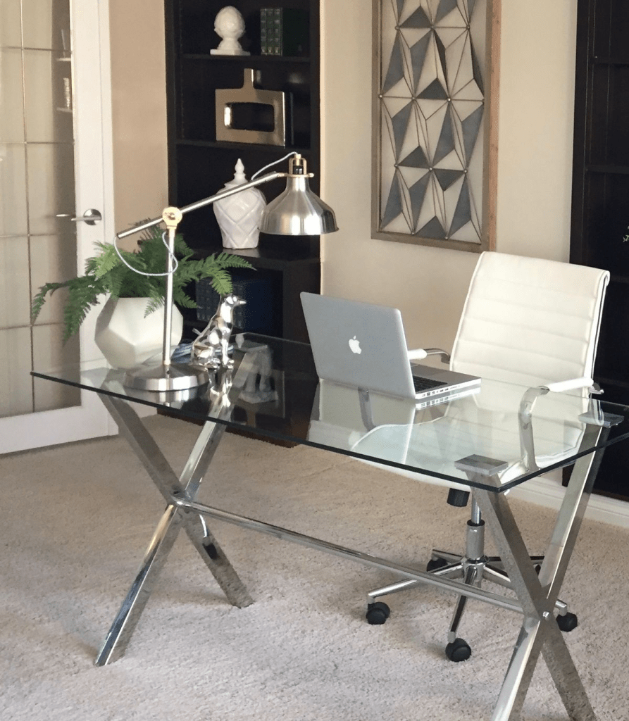 “Small home office design with glass computer desk, chrome legs, task lamp, indoor plant, and sleek modern decor in a neutral room.”