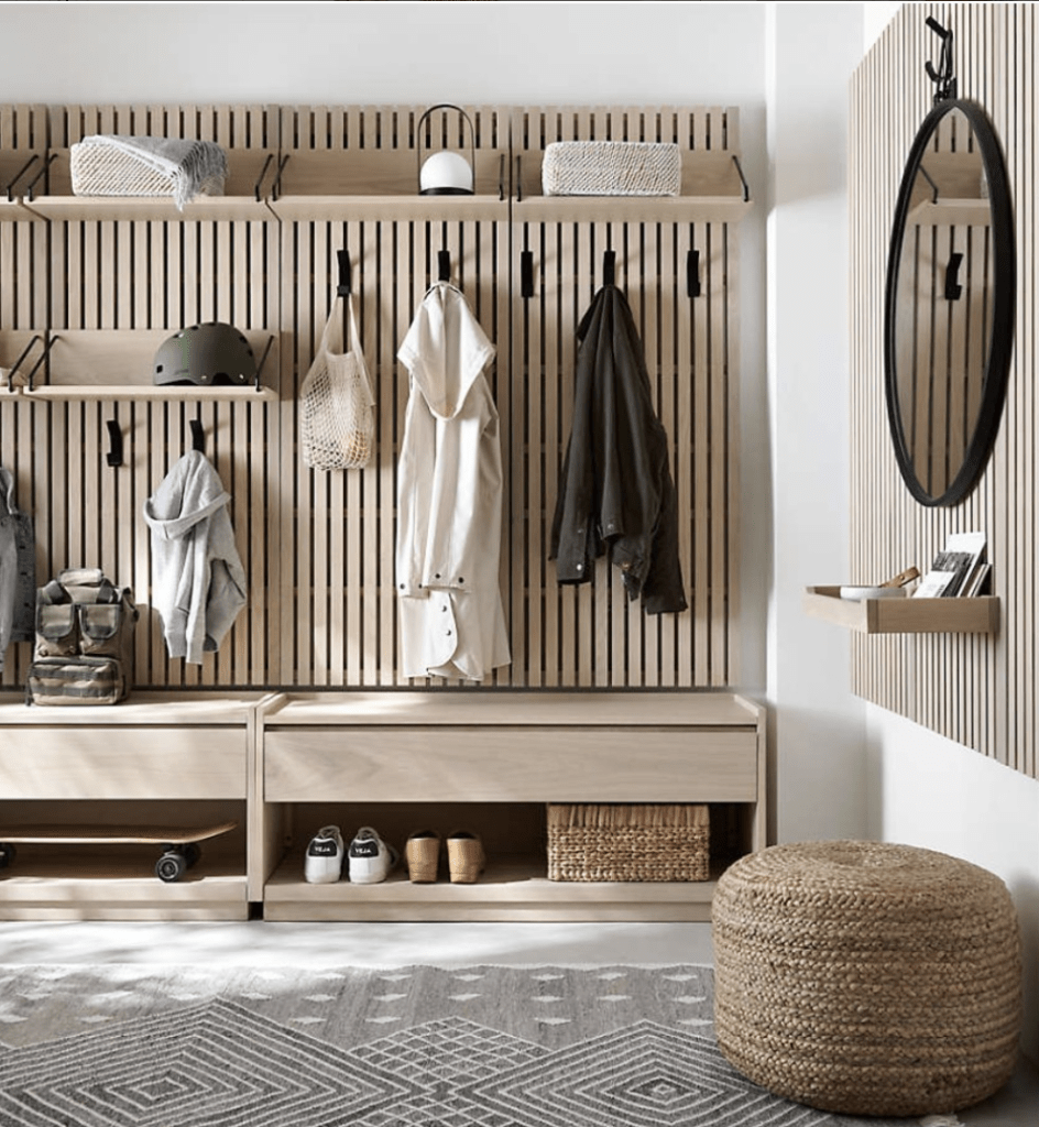 “Scandinavian mudroom entryway with slatted wood wall, floating shelves, black coat hooks, built-in bench storage, and minimalist decor.”