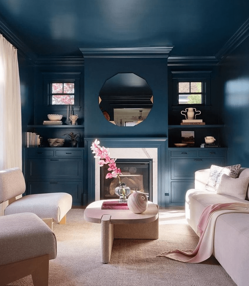 Deep blue living room with built-in shelves, white fireplace, octagon mirror, cream sofas, pink throw and orchid.