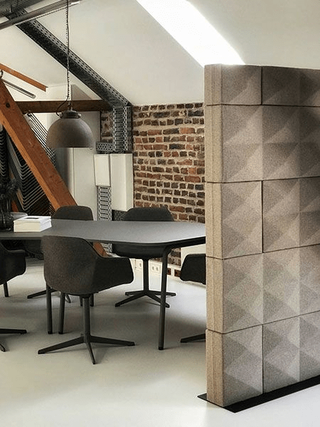 ontemporary workspace featuring a round meeting table, gray chairs, industrial brick and metal details, and a tall divider made of textured square panels.