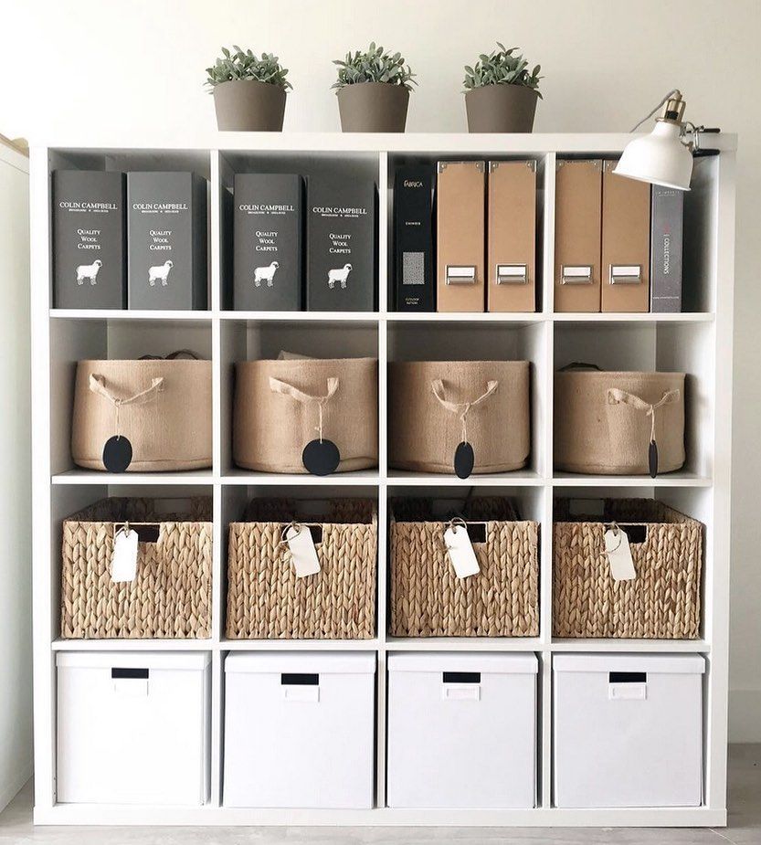 White cube storage shelf organized with binders. fabric bins. woven baskets. white boxes. and three small potted plants on top. The mix of open cubbies and labeled containers keeps office or home supplies sorted and easy to find.